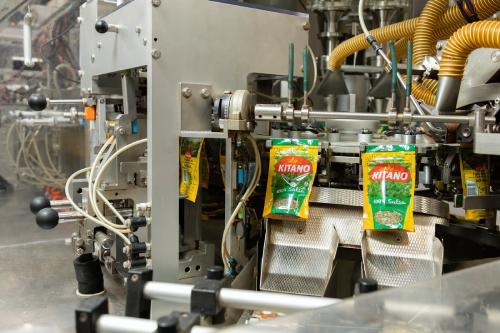 Packaging machine sealing Kitano spice packets on an assembly line in a factory setting.