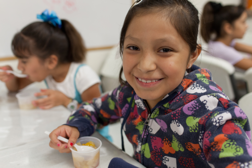 A smiling girl in a colorful hoodie holds a cup with food, while another girl eats in the background.