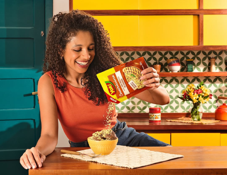 A woman in an orange top pours seasoning onto food in a yellow bowl, in a kitchen with colorful cabinets and decor.