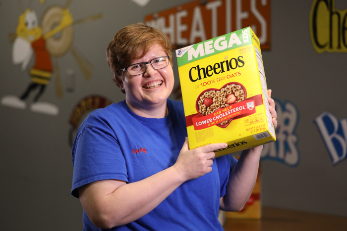 Person smiling while holding a large box of Cheerios cereal in front of a decorated wall.