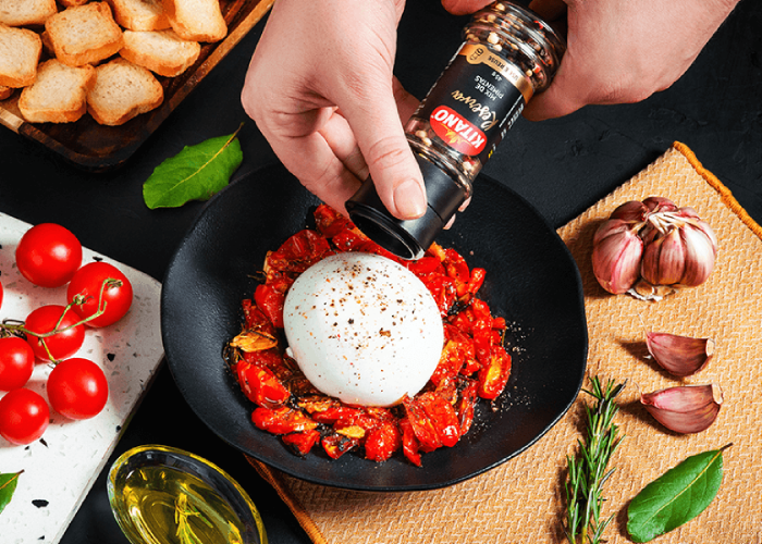 Hands seasoning a plate of burrata with cherry tomatoes. Surrounding items include bread, garlic, and herbs.