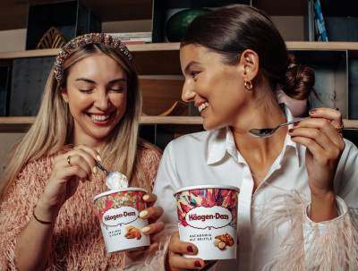 Two women seated, smiling, enjoying Häagen-Dazs ice cream from tubs with spoons, against a bookshelf backdrop.